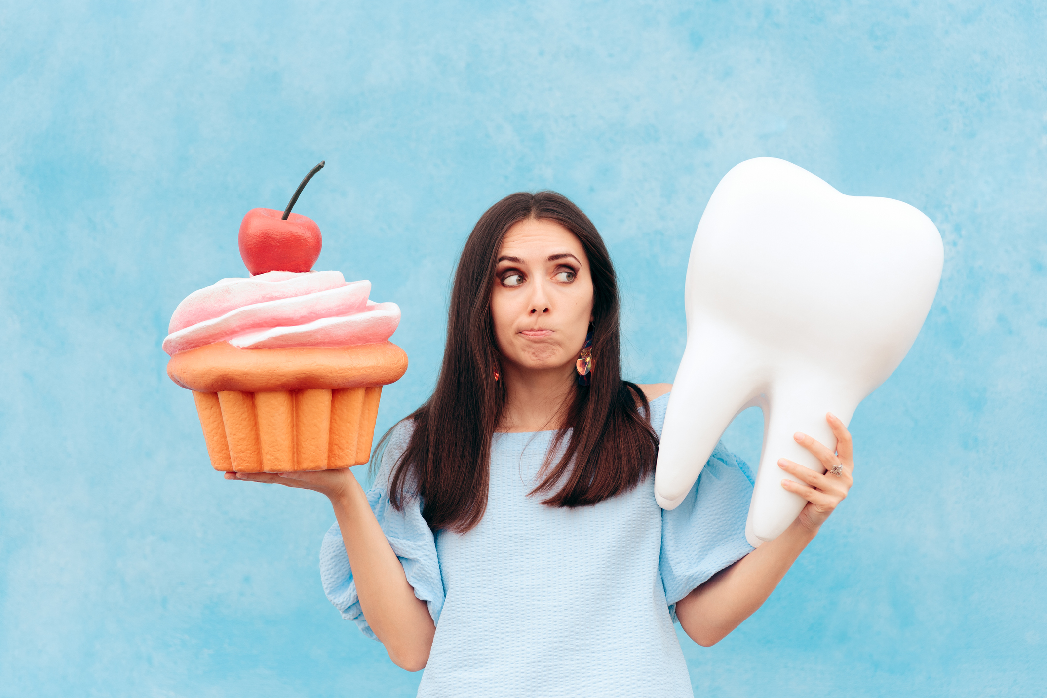 A woman with a conflicted expression holds a giant cupcake in one hand and a large model of a healthy white tooth in the other