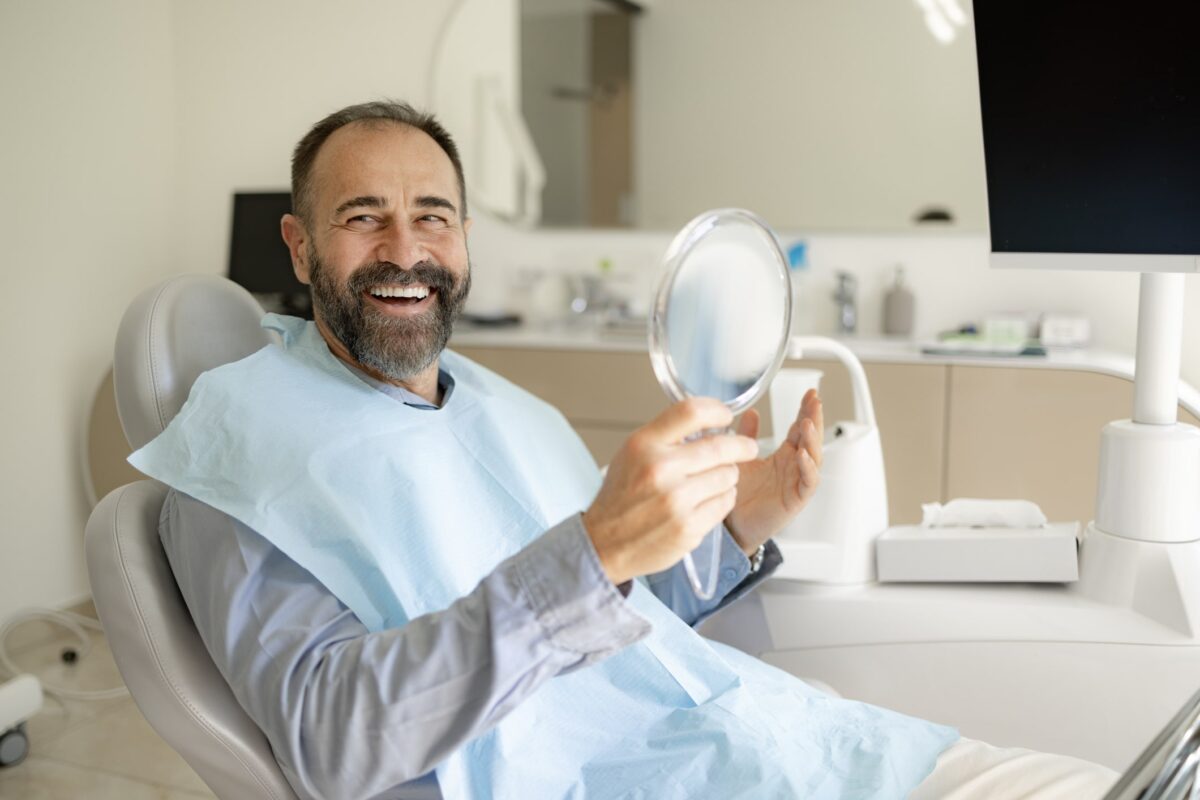 A happy man in a dental chair smiling widely while looking at his reflection in a handheld mirror