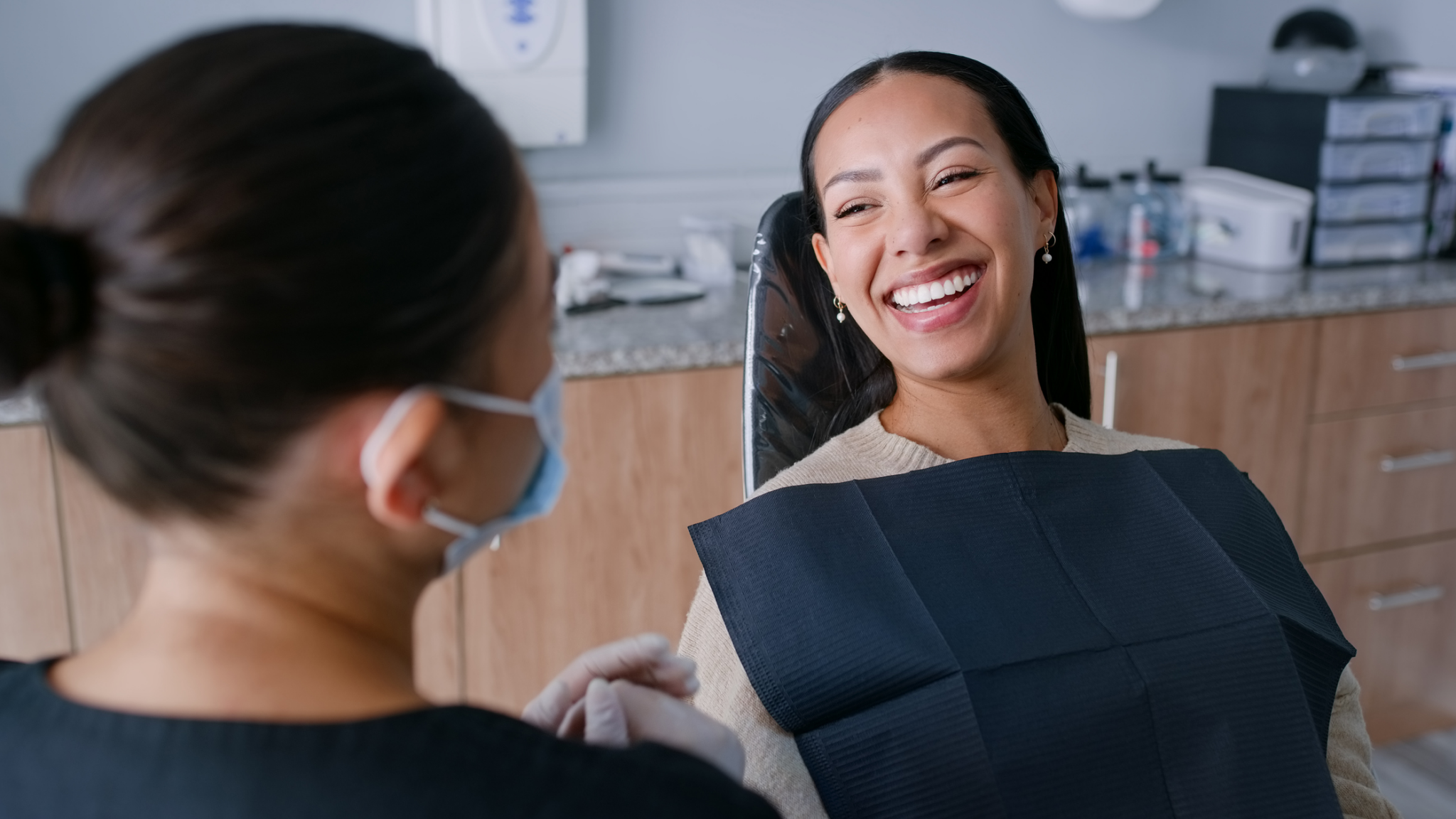 Woman smiling while seated in a reclined dental chair, with a sheet covering her neck and chest during treatment